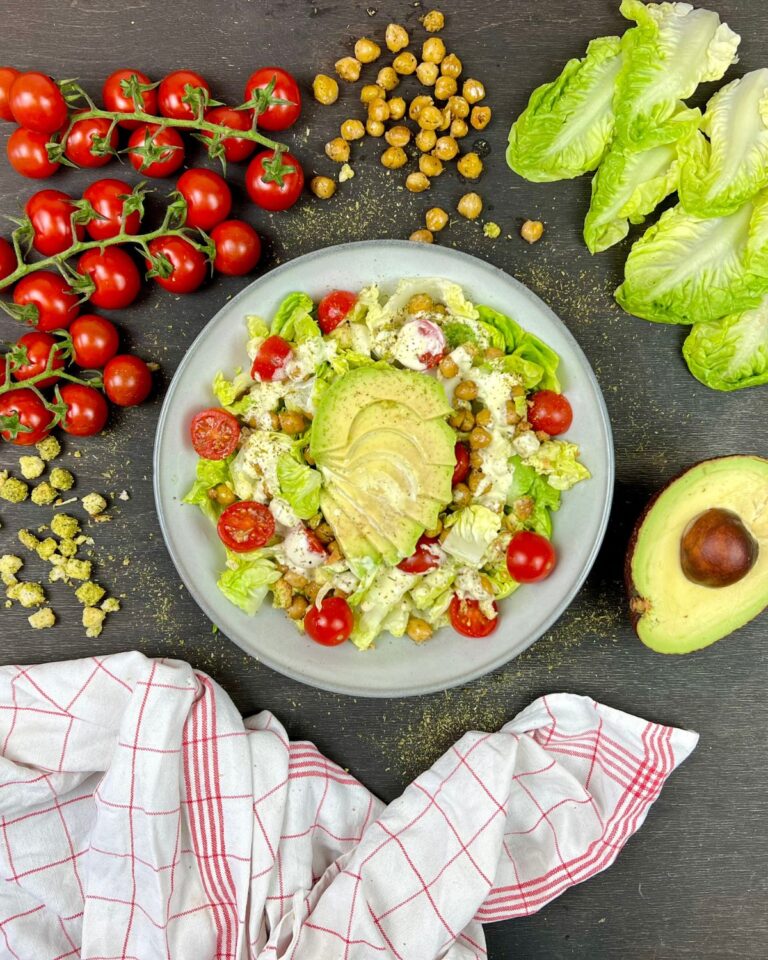 Ein vegetarischer Caesar Salad mit Salat, Cherrytomaten, Kichererbsen und Avocadoscheiben ruht auf einem grauen Teller. Um den Teller herum liegen Cherrytomaten am Strauch, ganze Kichererbsen, Romanasalatblätter, eine halbe reife Avocado und ein weiß-rot kariertes Tuch.