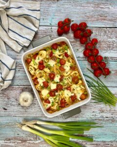 Ein gebackener vegetarischer Tortellini-Auflauf in einer weißen Auflaufform, belegt mit geschmolzenem Käse, Cherrytomaten und grünen Kräutern. Um die Form herum befinden sich Kirschtomatenbündel am Strauch, eine Knoblauchknolle, Frühlingszwiebeln, ein Bund Schnittlauch und ein gestreiftes Küchentuch auf einer rustikalen Holzoberfläche.