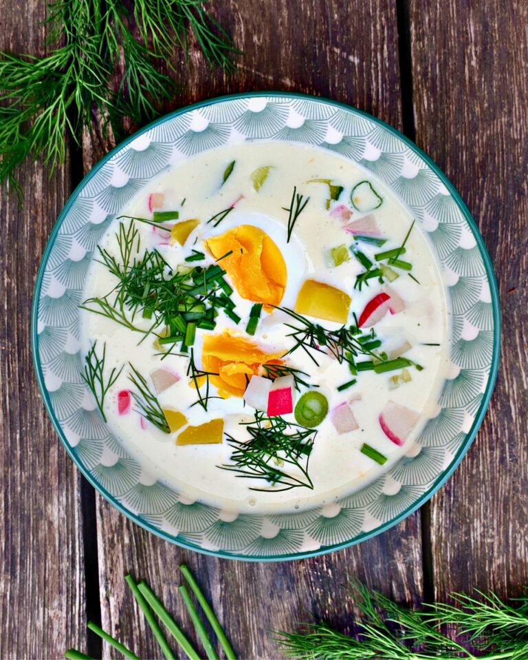 Eine Schüssel cremiges Vegetarisches Okroschka mit geschnittenen Eiern, Radieschen, Kartoffeln, Frühlingszwiebeln und frischem Dill steht auf einem Holztisch.