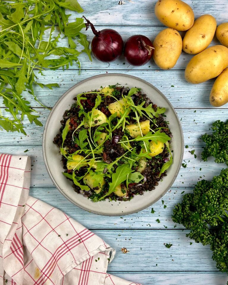 Ein Teller mit gemischtem Salat aus Rucola, Zwiebeln, Linsen und gegrillten Kartoffeln steht auf einer Holzoberfläche. Um den Teller herum liegen frischer Rucola, rote Zwiebeln, Kartoffeln und Petersilie. Am Rand des Tisches hängt ein rot-weiß kariertes Tuch, auf dem ein farbenfroher Linsen-Kartoffelsalat liegt.